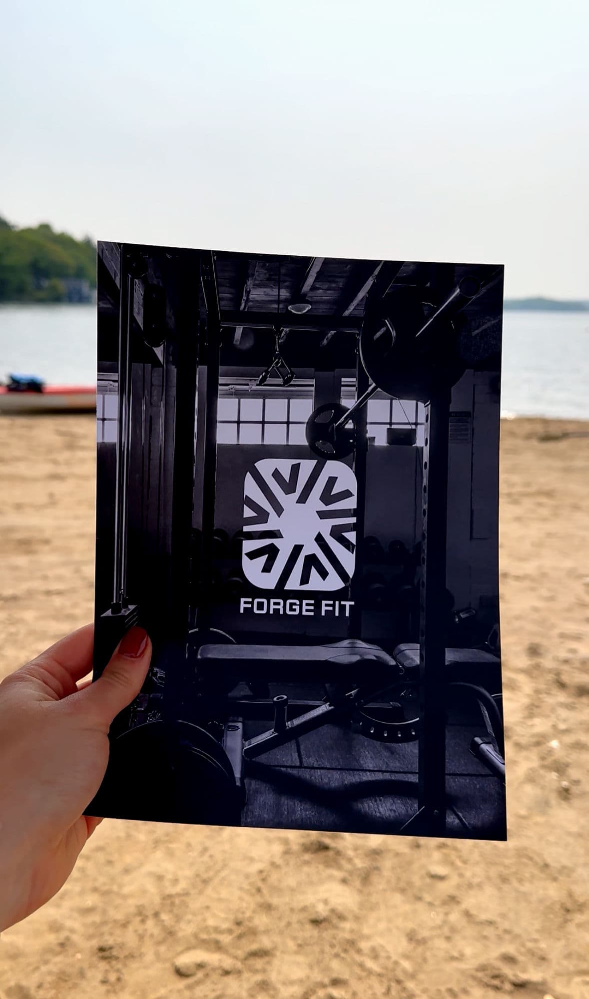 Hand holding a Forge Fit print with sunburst logo over a gym photograph at the beach